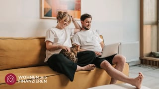 Junges Paar auf Couch nutzt Baufinanzierungsrechner an Laptop.